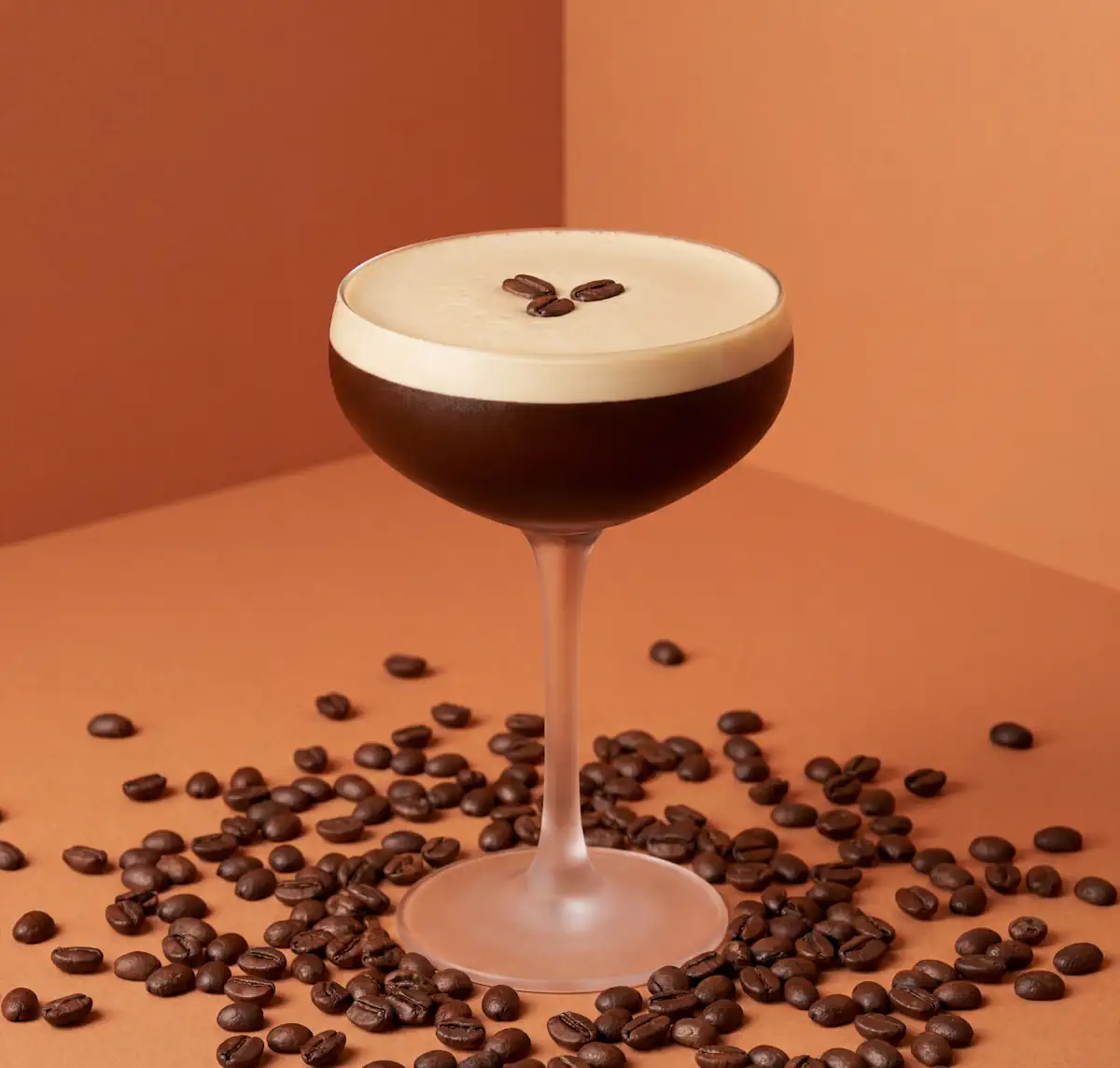 Espresso Martini cocktail with a thick, creamy nitro foam head and three coffee beans as garnish, presented in a coupe glass against a warm amber and dark brown background, serving from a Espresso Martini machine Naiko within 4 seconds, the worlds fastest Espresso Martini.