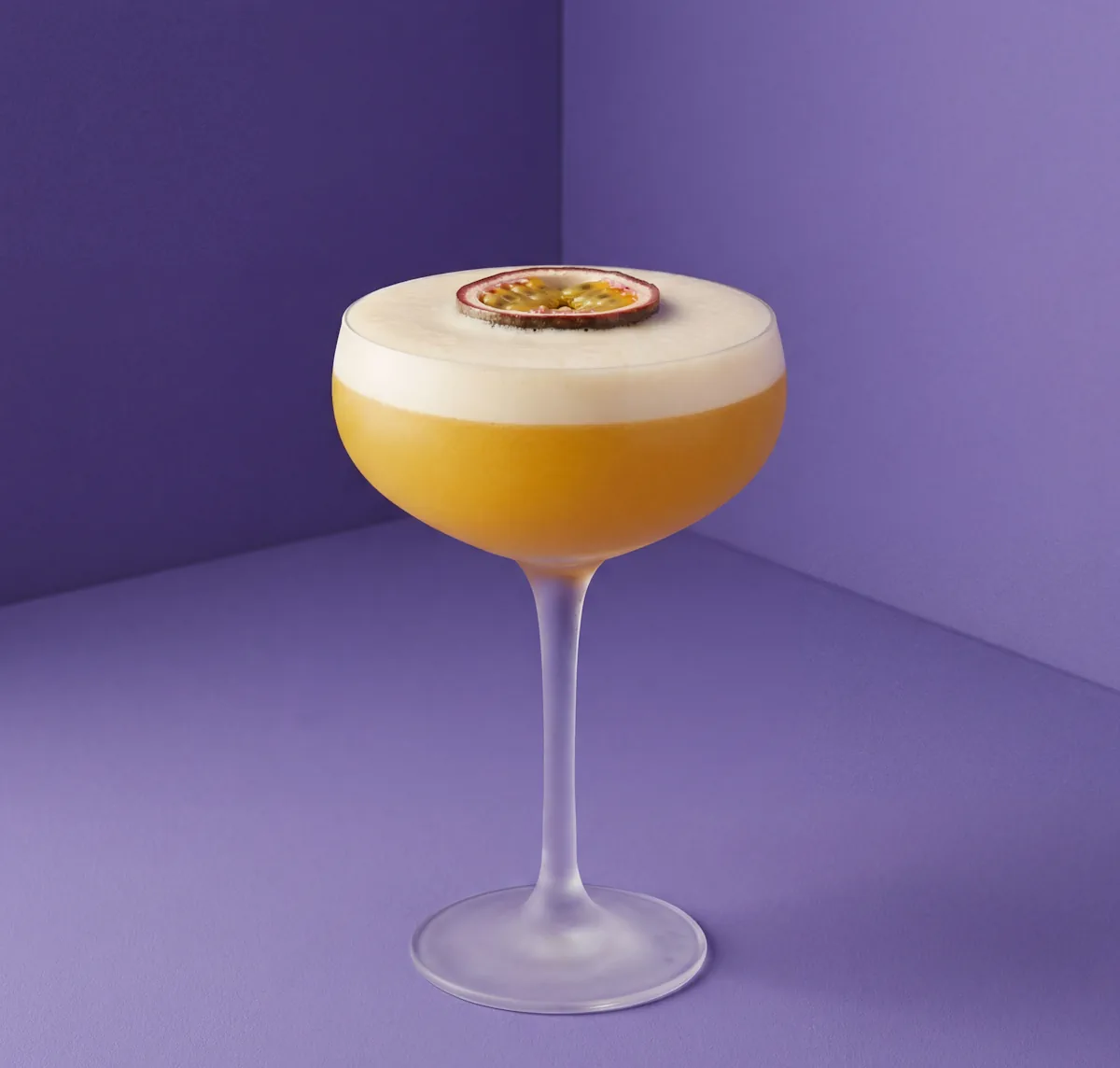 Pornstar Martini cocktail with vanilla vodka and passion fruit, topped with a thick white foam crown and a half passion fruit garnish, set against a vibrant purple studio background.